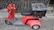 A red three-wheeled scooter with a large, weathered blue and brown storage box attached to the back. The scooter has a basket on the front, handlebars, and a cushioned seat. It is parked on a textured street surface, adjacent to a wooden fence and stone pavement.