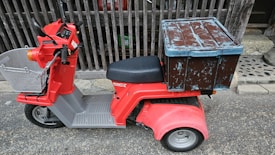 A red three-wheeled scooter with a large, weathered blue and brown storage box attached to the back. The scooter has a basket on the front, handlebars, and a cushioned seat. It is parked on a textured street surface, adjacent to a wooden fence and stone pavement.