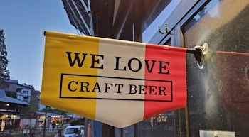 A rectangular flag with yellow, white, and red vertical stripes hangs on a glass surface. The text 'WE LOVE CRAFT BEER' is printed in bold, black letters. The background shows a blurred street scene with buildings and trees.
