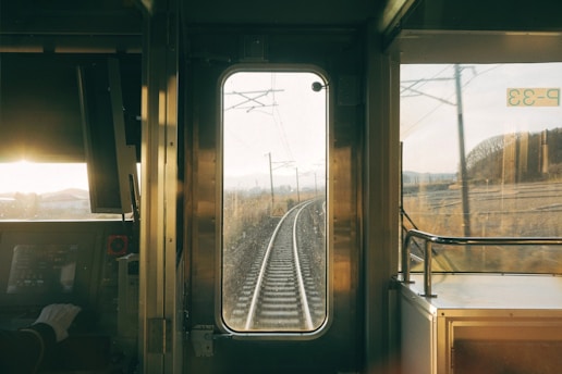 The interior view of a train's cockpit shows a railway track extending into the distance through a large central window. Sunlight filters through, casting a warm glow onto the control panel. A hand rests nearby, possibly of a train conductor. The landscape outside appears barren and slightly wooded in the background.