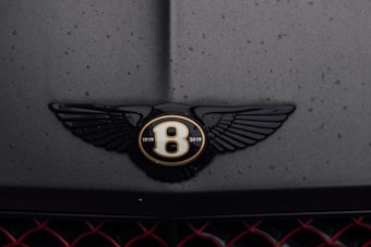 A close-up of a car emblem featuring a black winged logo with gold accents and the years 1919 to 2019 inscribed. The logo is on a dark, matte surface that has water droplets on it. Below the emblem, the car grill with a red mesh pattern is partially visible.