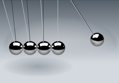 Five metallic spheres are suspended from a frame in a row on thin strings. Four of the spheres are still and touching, while the fifth sphere on the right is pulled away to the side, creating the motion of a Newton's cradle.