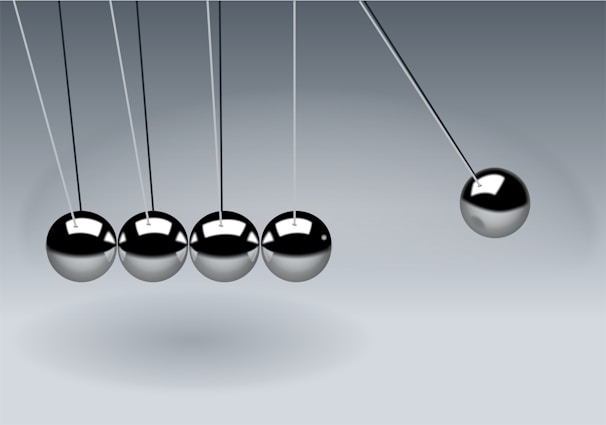 Five metallic spheres are suspended from a frame in a row on thin strings. Four of the spheres are still and touching, while the fifth sphere on the right is pulled away to the side, creating the motion of a Newton's cradle.