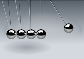 Five metallic spheres are suspended from a frame in a row on thin strings. Four of the spheres are still and touching, while the fifth sphere on the right is pulled away to the side, creating the motion of a Newton's cradle.