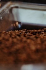 A close-up view of roasted coffee beans with a shallow depth of field. The beans appear rich in texture and are held in what seems to be a transparent container. The background is slightly blurred, emphasizing the focus on the roasted beans.