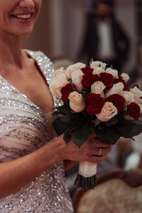 A person is holding a bouquet of roses, featuring red and light pink flowers. The attire is adorned with sparkling embellishments, suggesting formal or celebratory wear. The setting appears elegant, with blurred background elements, including a person in formal attire, hinting at a possible event or gathering.