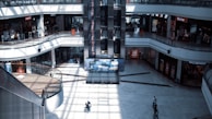 A spacious multi-level shopping mall with several retail stores, including Levi's and Flormar. The central feature is a large elevator surrounded by glass panels. Sunlight streams in through the high windows, casting geometric shadows on the floor. A few people are scattered across the open area, some walking and others standing.