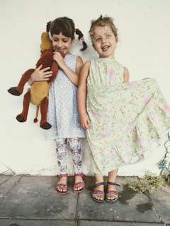 Two young children are standing against a light-colored wall, both smiling. One child is holding a plush toy, while the other is lifting the skirt of her floral dress playfully. They are wearing colorful dresses, patterned leggings, and sandals.