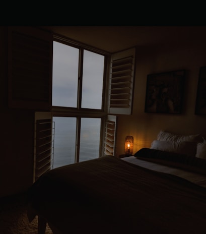 A peaceful bedroom with white linens, wooden accents, and a large window showing the sea and morning light.