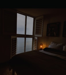 A dimly lit bedroom features a large window with shutters, revealing an overcast sky and a view of the ocean. The room is minimally furnished, including a bed with multiple pillows and a small, glowing lamp on a side table. Dark tones dominate, creating a cozy and serene atmosphere.