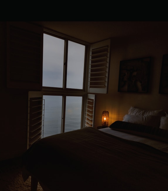 A dimly lit bedroom features a large window with shutters, revealing an overcast sky and a view of the ocean. The room is minimally furnished, including a bed with multiple pillows and a small, glowing lamp on a side table. Dark tones dominate, creating a cozy and serene atmosphere.