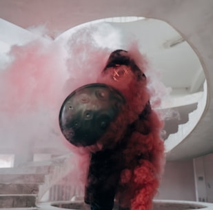 A person is obscured by vibrant pink smoke while holding a circular shield, standing on a spiral staircase. The environment appears to be indoors with concrete structures.