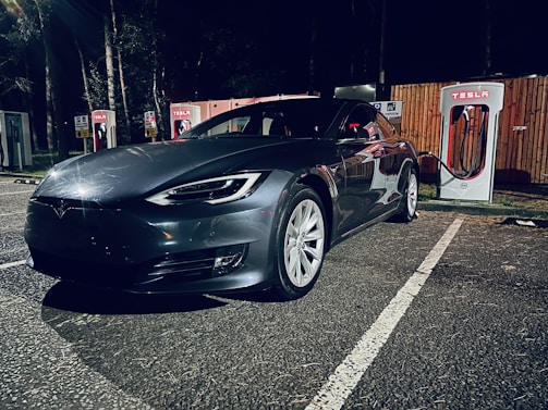 A sleek, dark-colored Tesla car is parked in an outdoor area at night. The car is positioned in front of a series of Tesla charging stations, one of which is actively charging the vehicle. The setting is dimly lit, with surrounding trees and a wooden fence in the background.