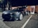 A sleek, dark-colored Tesla car is parked in an outdoor area at night. The car is positioned in front of a series of Tesla charging stations, one of which is actively charging the vehicle. The setting is dimly lit, with surrounding trees and a wooden fence in the background.