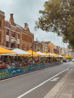A lively street scene with outdoor seating under yellow umbrellas along a sidewalk. People are sitting and dining in a row of cafes or restaurants, bordered by artistic barriers. The architecture features historical buildings with brown and red bricks, contrasted by a large tree on the opposite side of the road.