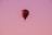 A hot air balloon with a checkered pattern of red, black, and white colors floats gently in the sky. The background is a gradient of soft pink and purple hues, suggesting a serene evening or early morning setting.