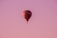 A hot air balloon with a checkered pattern of red, black, and white colors floats gently in the sky. The background is a gradient of soft pink and purple hues, suggesting a serene evening or early morning setting.