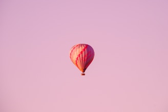 A colorful hot air balloon floats peacefully in a vast pink sky, capturing the serene beauty of flight and open spaces. The balloon is primarily red and white, featuring a classic stripe pattern.