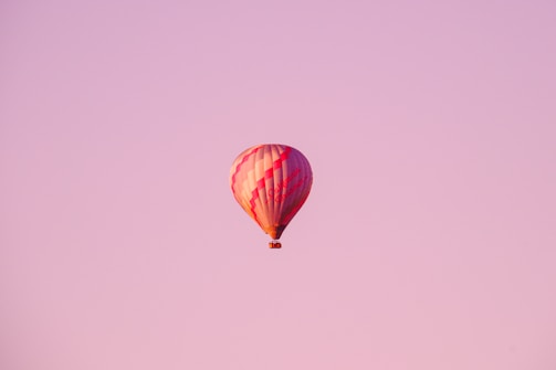 A colorful hot air balloon floats peacefully in a vast pink sky, capturing the serene beauty of flight and open spaces. The balloon is primarily red and white, featuring a classic stripe pattern.
