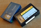 Two VHS-C video cassettes on a wooden surface. One is a Sony Premium 45 labeled case with blue coloring, and the other is a Fuji High Quality Plus HO+ EC-45 with a black casing and yellow label.