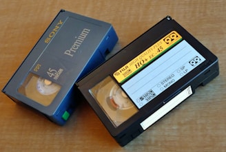 Two VHS-C video cassettes on a wooden surface. One is a Sony Premium 45 labeled case with blue coloring, and the other is a Fuji High Quality Plus HO+ EC-45 with a black casing and yellow label.