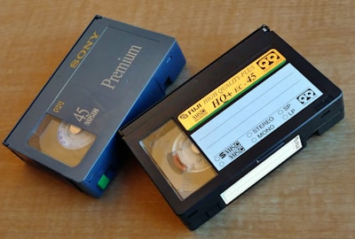 Two VHS-C video cassettes on a wooden surface. One is a Sony Premium 45 labeled case with blue coloring, and the other is a Fuji High Quality Plus HO+ EC-45 with a black casing and yellow label.