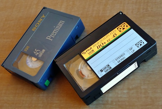 Two VHS-C video cassettes on a wooden surface. One is a Sony Premium 45 labeled case with blue coloring, and the other is a Fuji High Quality Plus HO+ EC-45 with a black casing and yellow label.