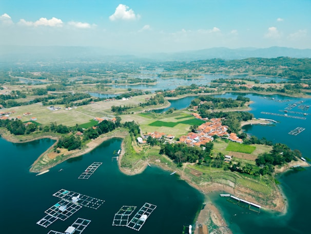 Aerial view of a rural landscape featuring a small village surrounded by lush greenery and bodies of water. The village consists of red-roofed houses clustered together. The water is calm with floating structures scattered across it, likely for aquaculture. Fields and patches of forested areas are visible, providing a mix of natural and cultivated scenery.