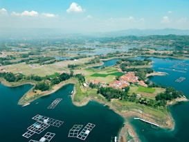 Aerial view of a rural landscape featuring a small village surrounded by lush greenery and bodies of water. The village consists of red-roofed houses clustered together. The water is calm with floating structures scattered across it, likely for aquaculture. Fields and patches of forested areas are visible, providing a mix of natural and cultivated scenery.