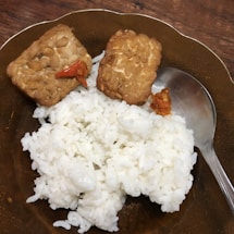 A plate holds a serving of white rice accompanied by two pieces of fried tempeh, each topped with a bit of red chili. A metal spoon rests on the plate's edge.
