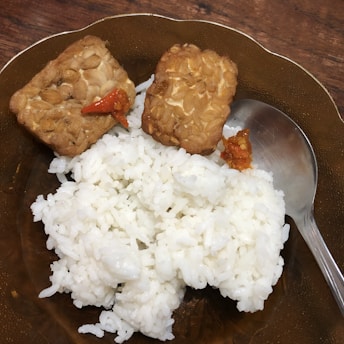 A plate holds a serving of white rice accompanied by two pieces of fried tempeh, each topped with a bit of red chili. A metal spoon rests on the plate's edge.