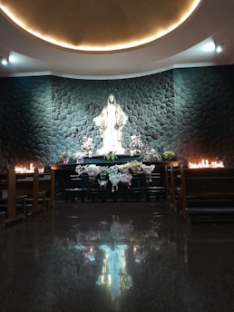 A serene chapel setting featuring a prominent statue of a religious figure surrounded by stone walls. The figure is depicted with open arms, with bouquets of flowers and lit candles placed artistically around the altar. The dim lighting adds a reflective and peaceful ambiance, enhanced by the polished floor capturing the reflection of the statue.