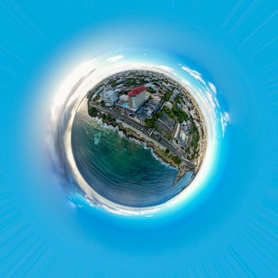 A 360-degree aerial view of a coastal city with buildings, roads, and greenery encircled by the ocean. The arrangement creates a petite planet effect with a bright blue sky surrounding the top.