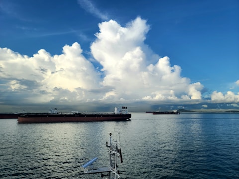 Large cargo ships float on calm waters under a vast, blue sky filled with fluffy, white clouds. The horizon is visible in the distance, with a faint outline of land.
