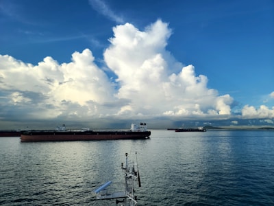 Large cargo ships float on calm waters under a vast, blue sky filled with fluffy, white clouds. The horizon is visible in the distance, with a faint outline of land.