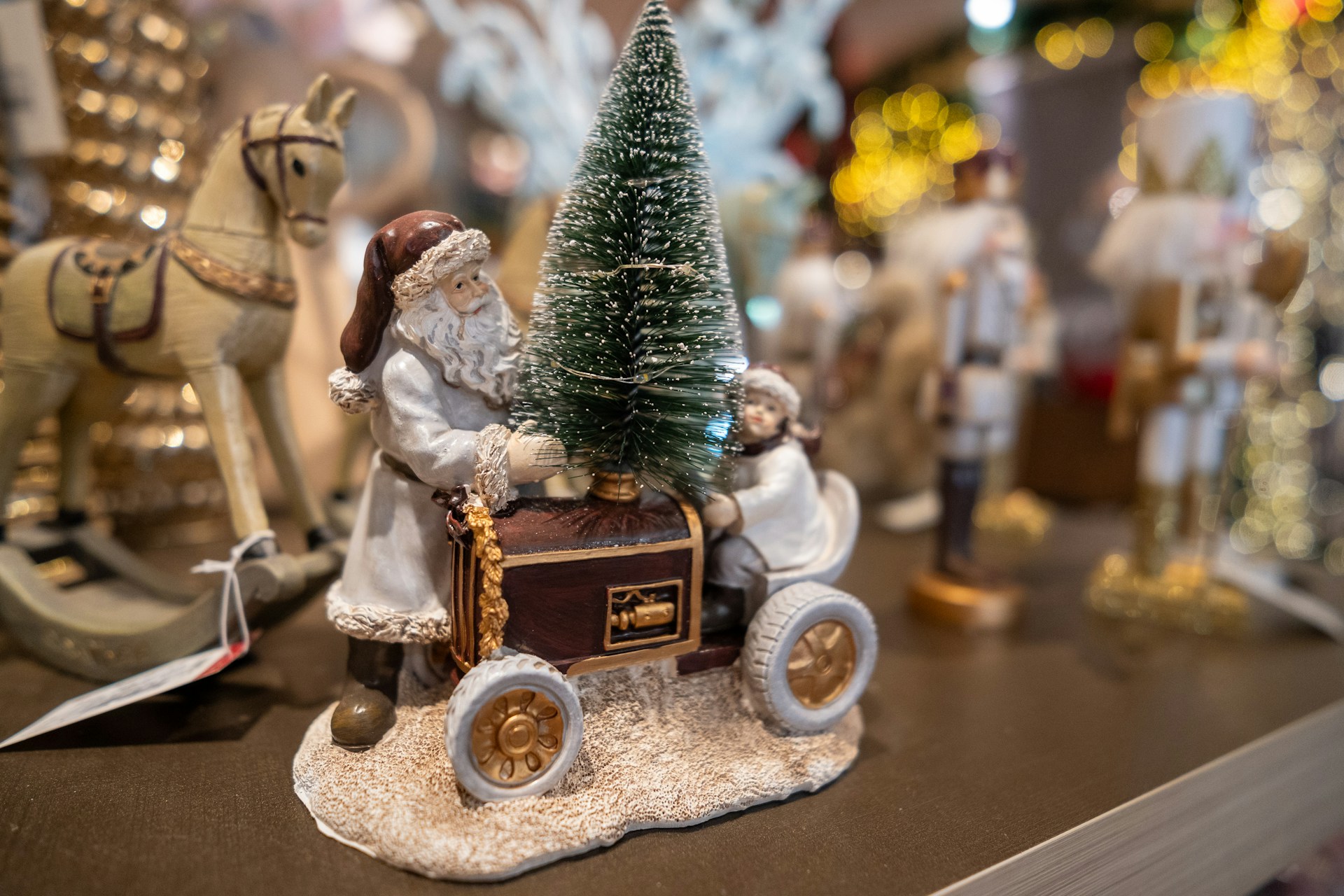 A festive scene with a decorative figure of Santa Claus pushing a cart carrying a small Christmas tree. Beside him, there's a child in winter clothing. In the background, blurred decorations and bright, glowing lights create a holiday ambiance. A wooden rocking horse is visible to the left.