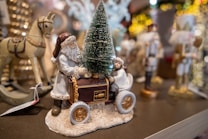 A festive scene with a decorative figure of Santa Claus pushing a cart carrying a small Christmas tree. Beside him, there's a child in winter clothing. In the background, blurred decorations and bright, glowing lights create a holiday ambiance. A wooden rocking horse is visible to the left.