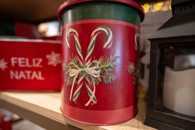 A decorative tin with a festive design featuring two candy canes crossed and adorned with greenery and a ribbon. The tin is prominently red with accents of green and white. In the background, there is a sign or box with the words 'FELIZ NATAL' in white text, suggesting a Christmas theme. A black lantern is also partially visible to the right.