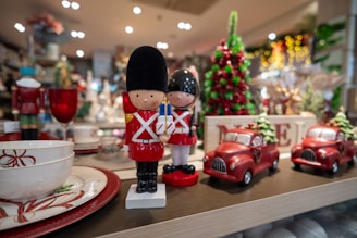 Two toy soldiers stand on a shelf alongside decorative red cars and festive dishware. In the background, a green and red Christmas tree decoration is visible, enhancing the holiday theme. The scene is set in a warmly lit store with an array of holiday ornaments and decorations.