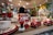Two toy soldiers stand on a shelf alongside decorative red cars and festive dishware. In the background, a green and red Christmas tree decoration is visible, enhancing the holiday theme. The scene is set in a warmly lit store with an array of holiday ornaments and decorations.