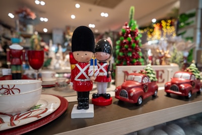 Two toy soldiers stand on a shelf alongside decorative red cars and festive dishware. In the background, a green and red Christmas tree decoration is visible, enhancing the holiday theme. The scene is set in a warmly lit store with an array of holiday ornaments and decorations.
