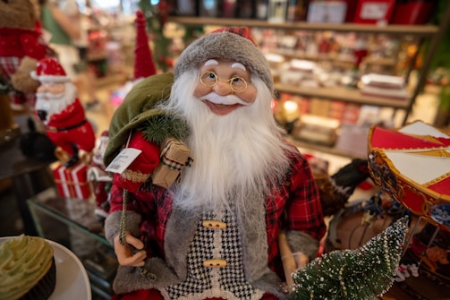 A detailed figurine of Santa Claus is displayed prominently. The Santa figurine is dressed in a traditional red and white outfit with a plaid pattern, complete with a Santa hat and wearing glasses. He carries a small sack and has a realistic, white beard and mustache. The background is filled with various festive decorations, including more Santa figurines, Christmas-themed items, and what appears to be a bakery with cupcakes visible in the foreground.