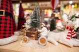 A crafted holiday scene featuring a figurine of a child in winter clothing on a small wooden vehicle, holding a Christmas tree. A plush reindeer with a red and white scarf stands nearby, and surrounding them are red and black checkered fabric decorations with gold buckles. The background has a warm festive ambiance with blurred lights and other holiday ornaments.