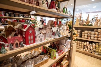 A store shelf filled with various Christmas-themed miniature houses and decorations. The foreground shows intricately designed houses in red, white, and natural wood tones, some with snow-like accents on the roofs. The background displays an assortment of holiday ornaments and boxed items, suggesting a festive shopping environment.