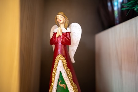 A decorative angel figurine dressed in a red robe with golden embellishments is positioned against a wooden backdrop. The angel has white wings and a serene expression, with hands clasped in a praying gesture.