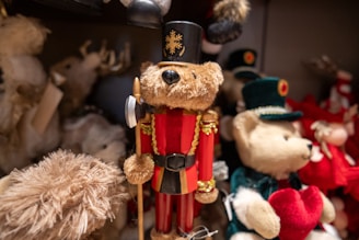 A collection of plush teddy bears with one prominently dressed as a soldier in red and gold uniform, complete with a tall black hat and holding a small axe. The surrounding bears are adorned in various outfits, with one holding a red heart. Soft textures and intricate details are visible, creating a festive and cozy atmosphere.