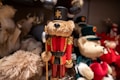 A collection of plush teddy bears with one prominently dressed as a soldier in red and gold uniform, complete with a tall black hat and holding a small axe. The surrounding bears are adorned in various outfits, with one holding a red heart. Soft textures and intricate details are visible, creating a festive and cozy atmosphere.