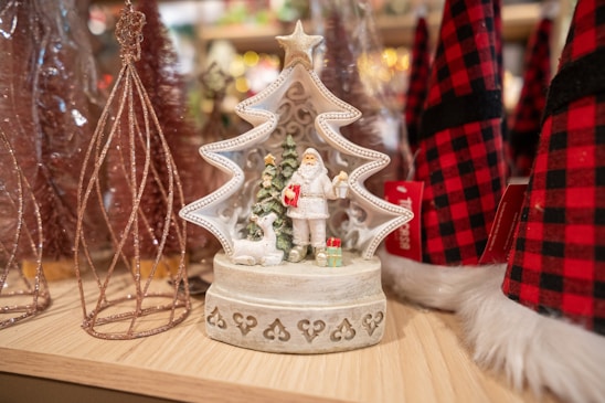 A decorative Christmas figurine featuring Santa Claus holding a lantern and standing beside a reindeer and a small Christmas tree. The figurine is shaped like a star-topped Christmas tree, placed on a wooden shelf surrounded by other festive decorations, including red and black plaid fabric hats and glittery wire trees.