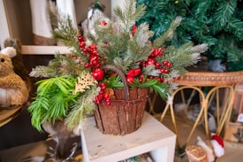 A rustic wooden basket adorned with pine branches, red berries, and red ribbon decorations is placed on a light wooden shelf. To the left, there's a decorative stuffed animal resembling a small woodland creature. In the background, a Christmas tree with green branches is partially visible.