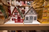 Two miniature wooden houses with a festive theme, featuring intricate details like fences and windows. The house on the left is predominantly white with red accents, including a red triangle pattern and matching fence. The house on the right is gray with a detailed cut-out design and a chimney, sitting beside golden gift boxes.